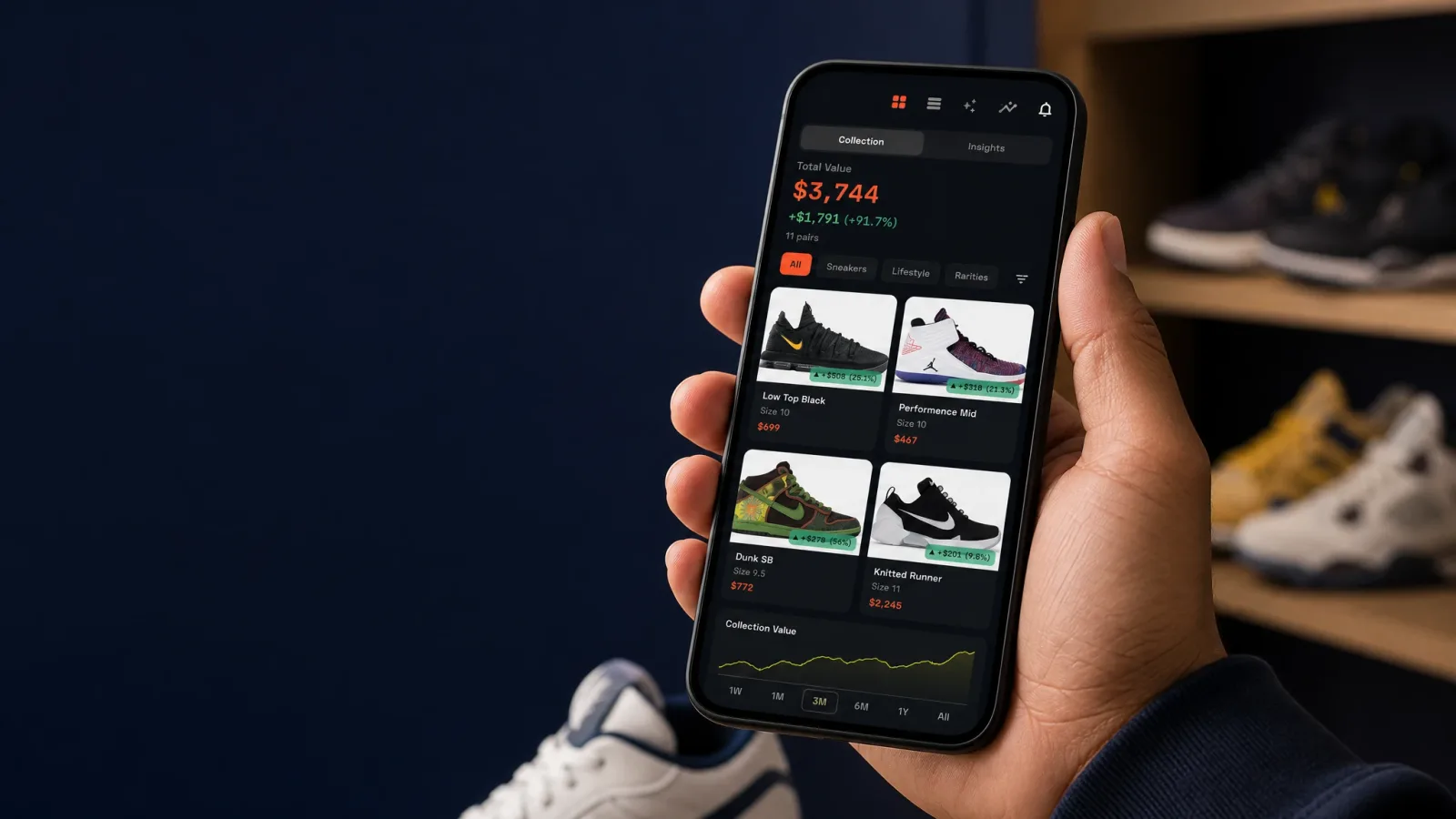 A close-up of a hand holding a smartphone displaying a clean sneaker portfolio app interface with shoe thumbnails, a collection value figure, and a chart, with a blurred sneaker shelf visible in the background.