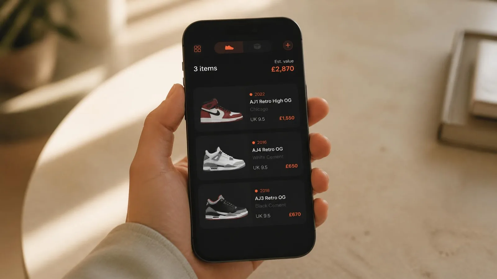 A person's hand holding a smartphone showing a dark-background sneaker collection tracking interface with value cards highlighted in orange, photographed in soft natural light against a warm background.