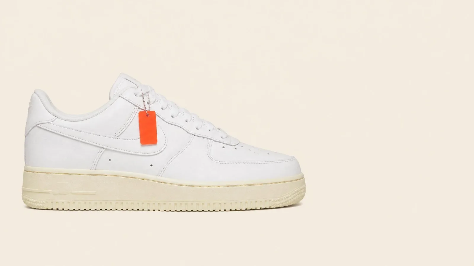 Dead stock white leather low-top sneaker in side-profile view against a warm off-white background, with a small orange authentication tag attached to the lace eyelet.