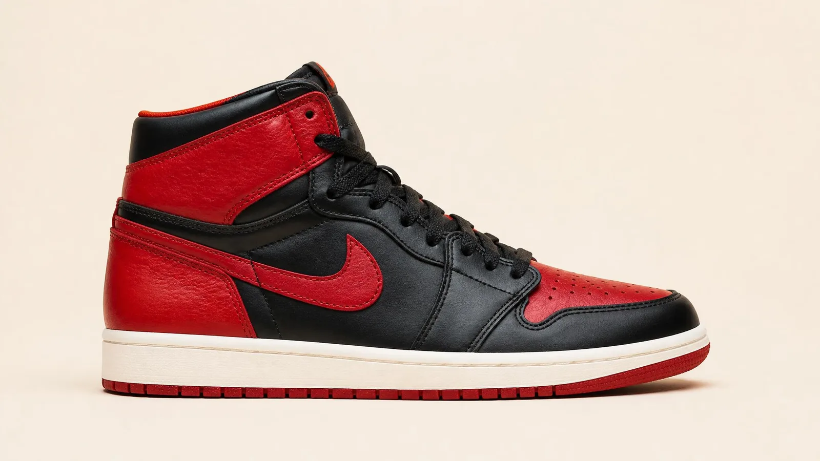 A side-profile studio shot of a pair of Air Jordan 1s in a classic red and black colourway, centred on a warm cream background with clean even lighting that highlights every panel and stitch detail.