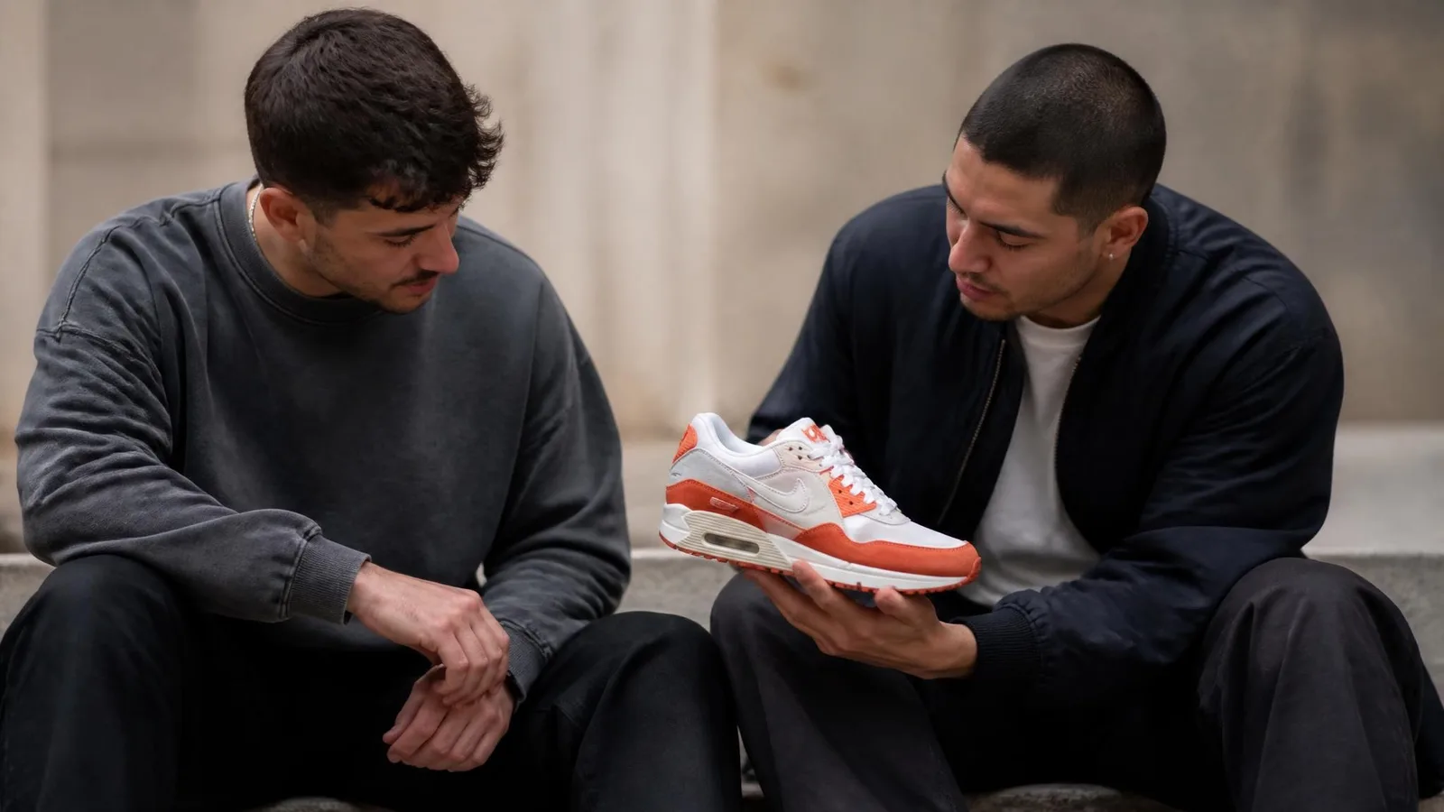 Two young men in casual streetwear sitting on a concrete step outdoors, leaning in to examine a sneaker with burnt-orange detailing held between them, shot in soft overcast daylight against a blurred urban background.