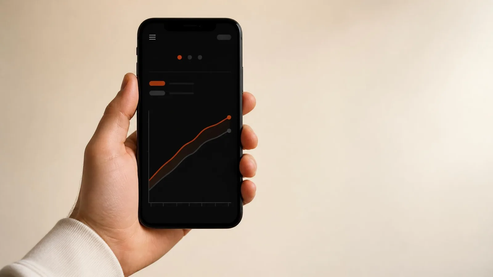 A hand holding a smartphone displaying a dark app screen with two performance chart lines, one in orange sitting above a benchmark line