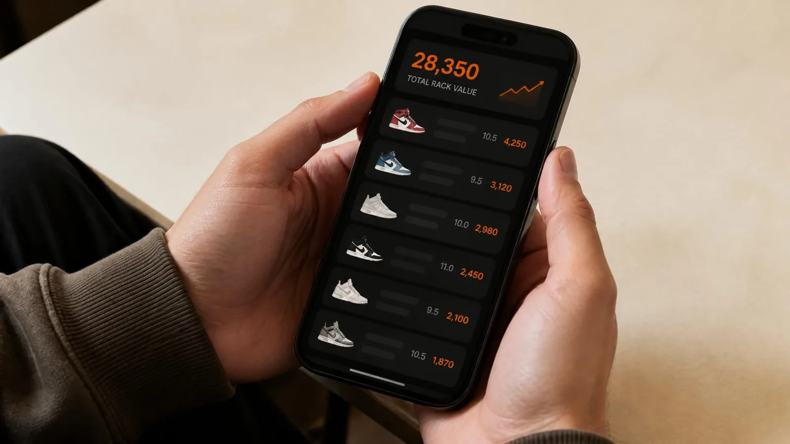 A collector's hands holding a smartphone showing a dark-background sneaker portfolio interface with pair names, sizes, and values listed and a burnt-orange total rack value highlighted at the top, resting above a warm off-white table surface.